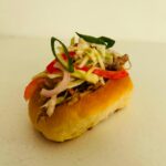 A delicious pulled pork sandwich topped with fresh cabbage, red peppers, and scallions, served on a soft bun against a minimalist background.
