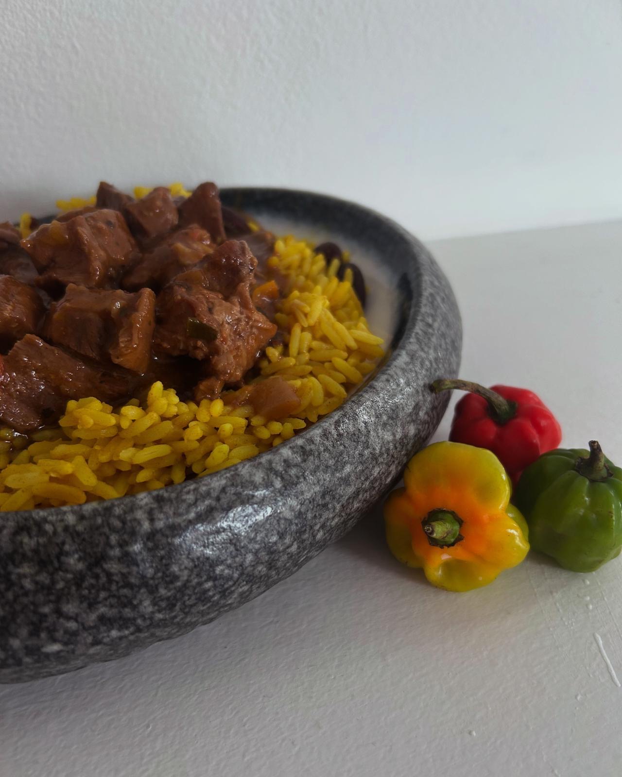 A gray bowl filled with yellow rice and chunks of beef, accompanied by colorful peppers on the side, against a plain white background.