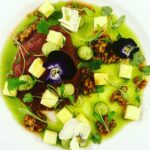 A beautifully arranged plate featuring a gourmet dish with seared meat, vibrant green sauce, edible flowers, diced vegetables, and garnishes, showcasing artistic culinary presentation.