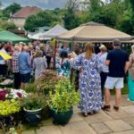 A bustling outdoor gathering with guests mingling under tents, surrounded by lush plants and flowers.
