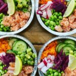 Four visually appealing salmon poke bowls with fresh vegetables, avocado, and lime, arranged on a wooden table.