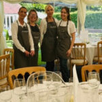 Four women dressed in aprons stand together in a beautifully arranged dining area with tables set for an event.