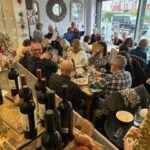 A lively dining scene featuring a group of people enjoying a meal together in a cozy atmosphere, with wine bottles on display and a festive setting.