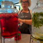 A woman in an apron, surprised, stands between two glass beverage dispensers filled with vibrant red and green drinks.