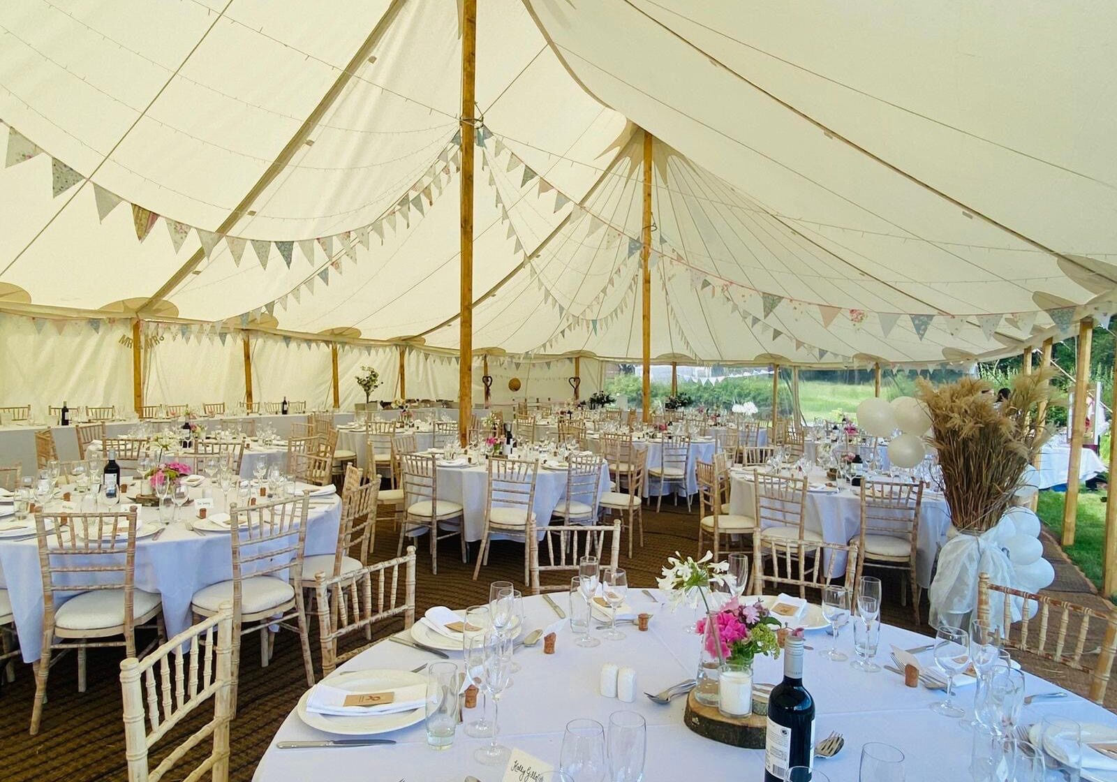 A beautifully arranged wedding reception tent featuring round tables with floral centerpieces, elegant chairs, and decorative bunting.